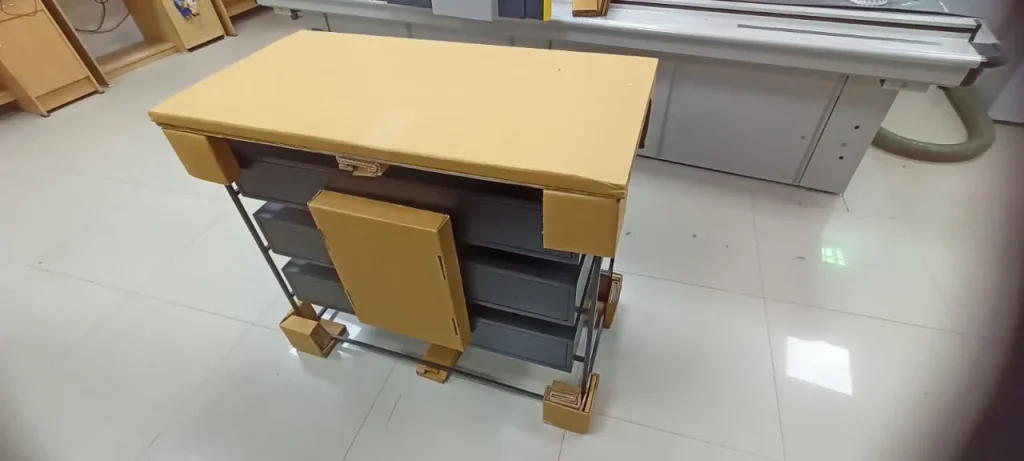 Metal-framed furniture unit with drawers protected by cardboard corner blocks, foot guards, a top cover panel and a front door panel