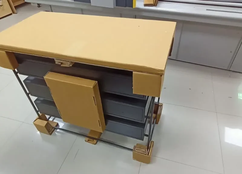 Metal-framed furniture unit with drawers protected by cardboard corner blocks, foot guards, a top cover panel and a front door panel